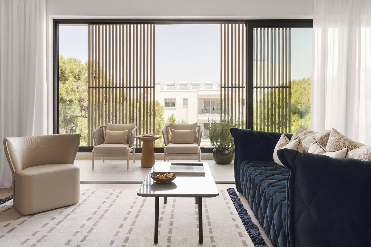 Een moderne woonkamer met een bank, twee fauteuils, een salontafel en een groot raam met jaloezieën.