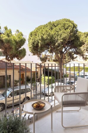 Un balcone con una sedia bianca, un tavolino con una ciotola di frutta e vista su auto parcheggiate e palazzi.
