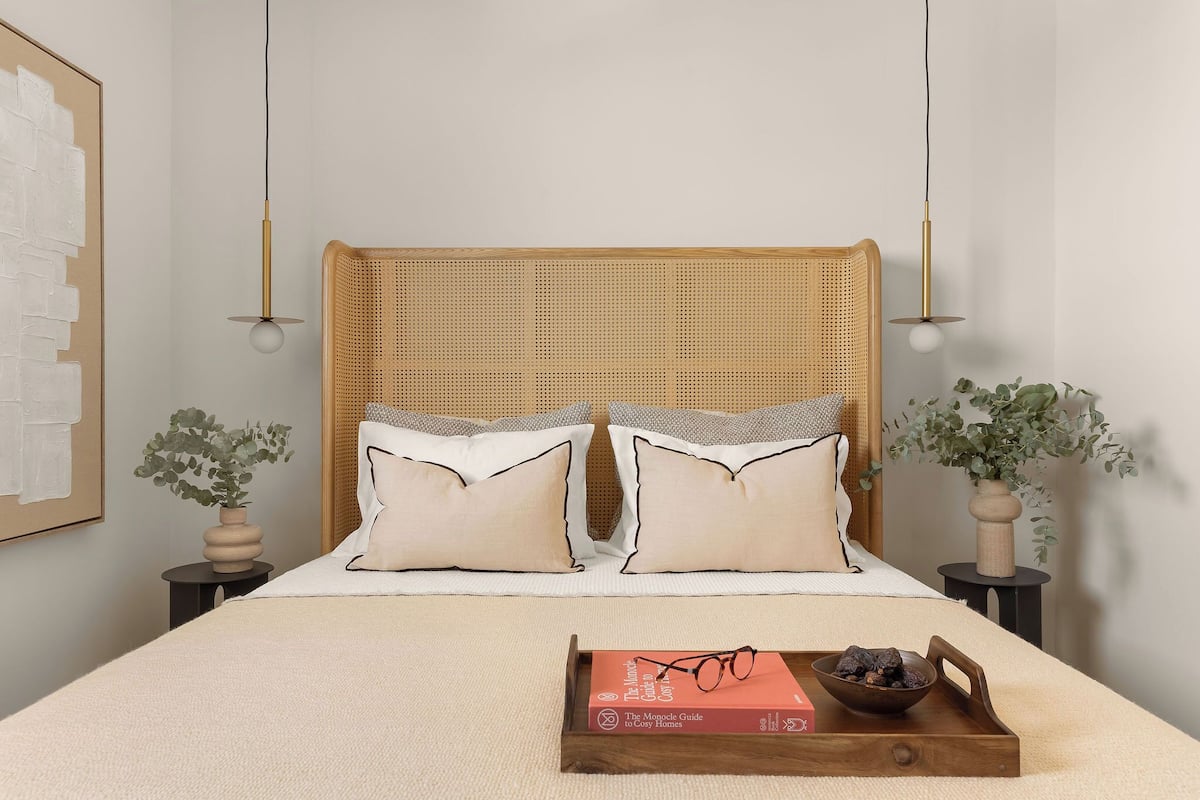 Een net opgemaakt bed met een geweven hoofdbord, twee nachtkastjes met lampen, een ingelijst kunstwerk aan de muur, en een dienblad met een boek en brillen op het bed.