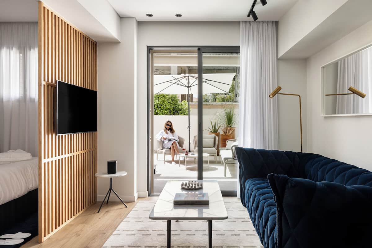 Een moderne hotelkamer met een bed, een televisie bevestigd aan een houten paneel, een donkerblauwe bank, een klein tafeltje met een boek en een glazen schuifdeur die leidt naar een buitenruimte met een stoel en een tafel.