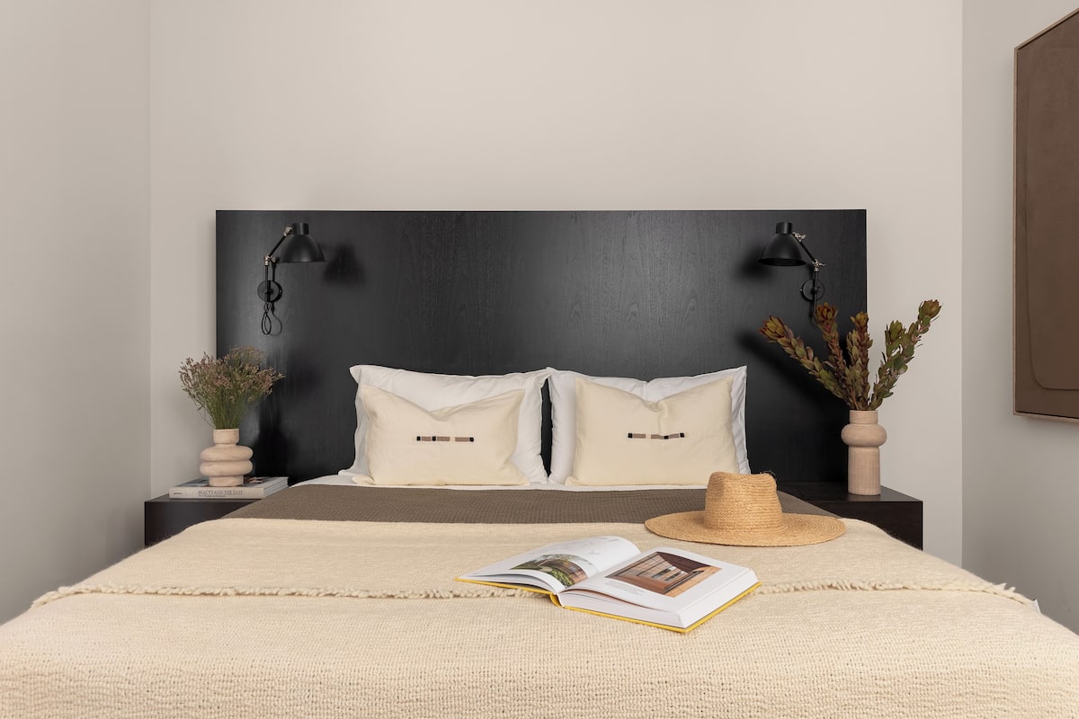 Een net opgemaakt bed met een donker hoofdbord, twee nachtkastjes met lampen, een boek en een strohoed.