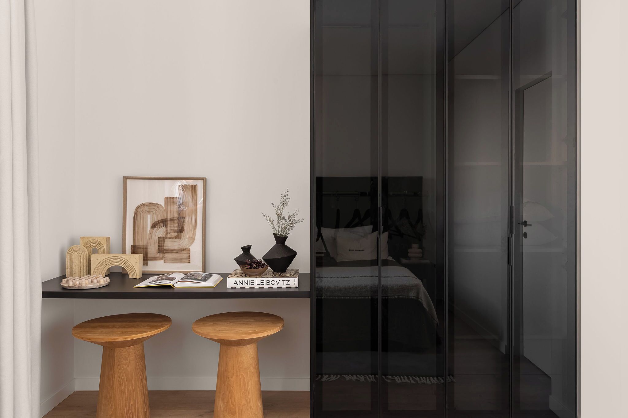 Una habitación moderna con una puerta de vidrio que da a un dormitorio. Hay un escritorio con un libro titulado 'ANNIE LEIBOVITZ' y dos taburetes de madera.