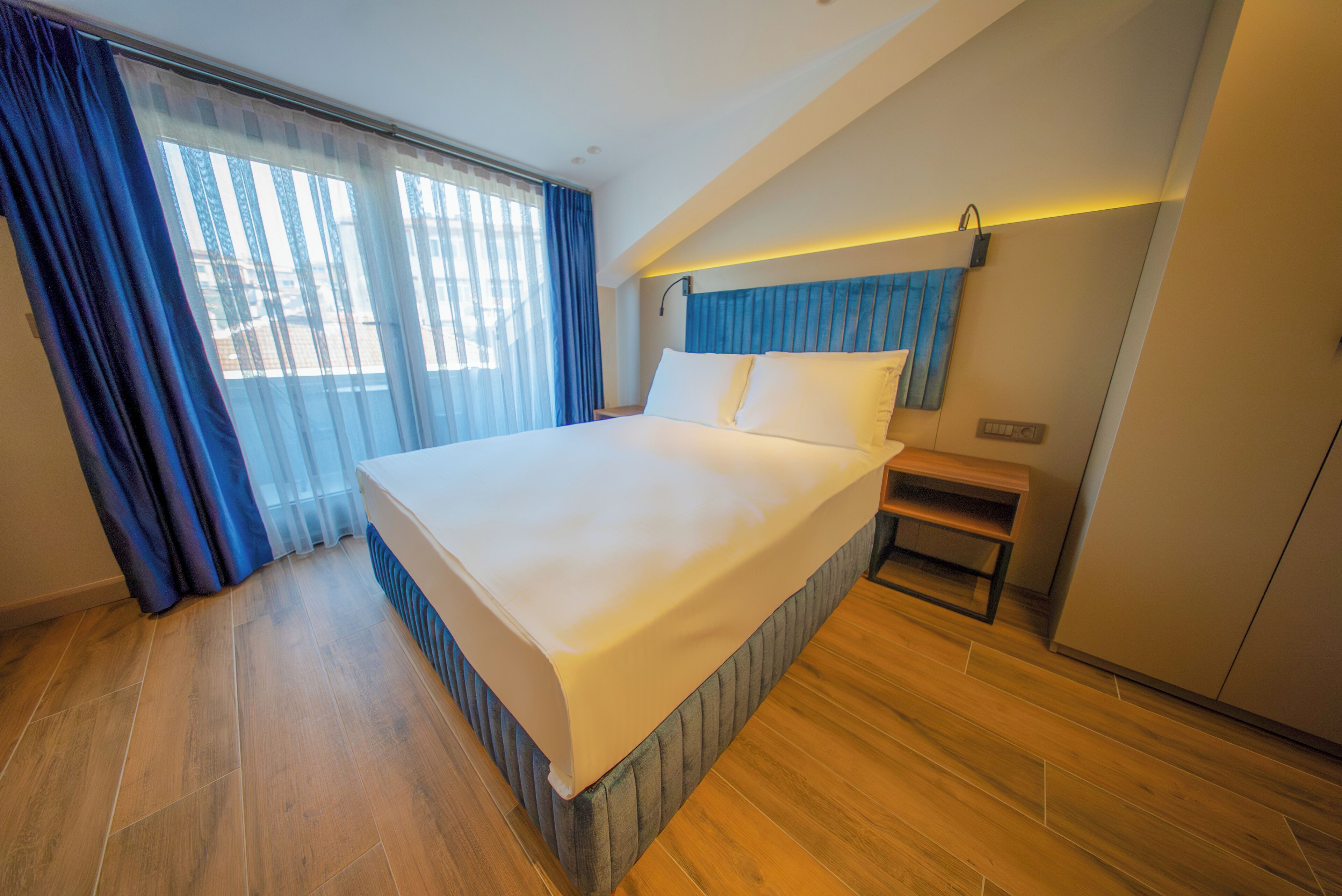 A modern hotel room with a large bed, wooden flooring, a nightstand, and a view through the window.