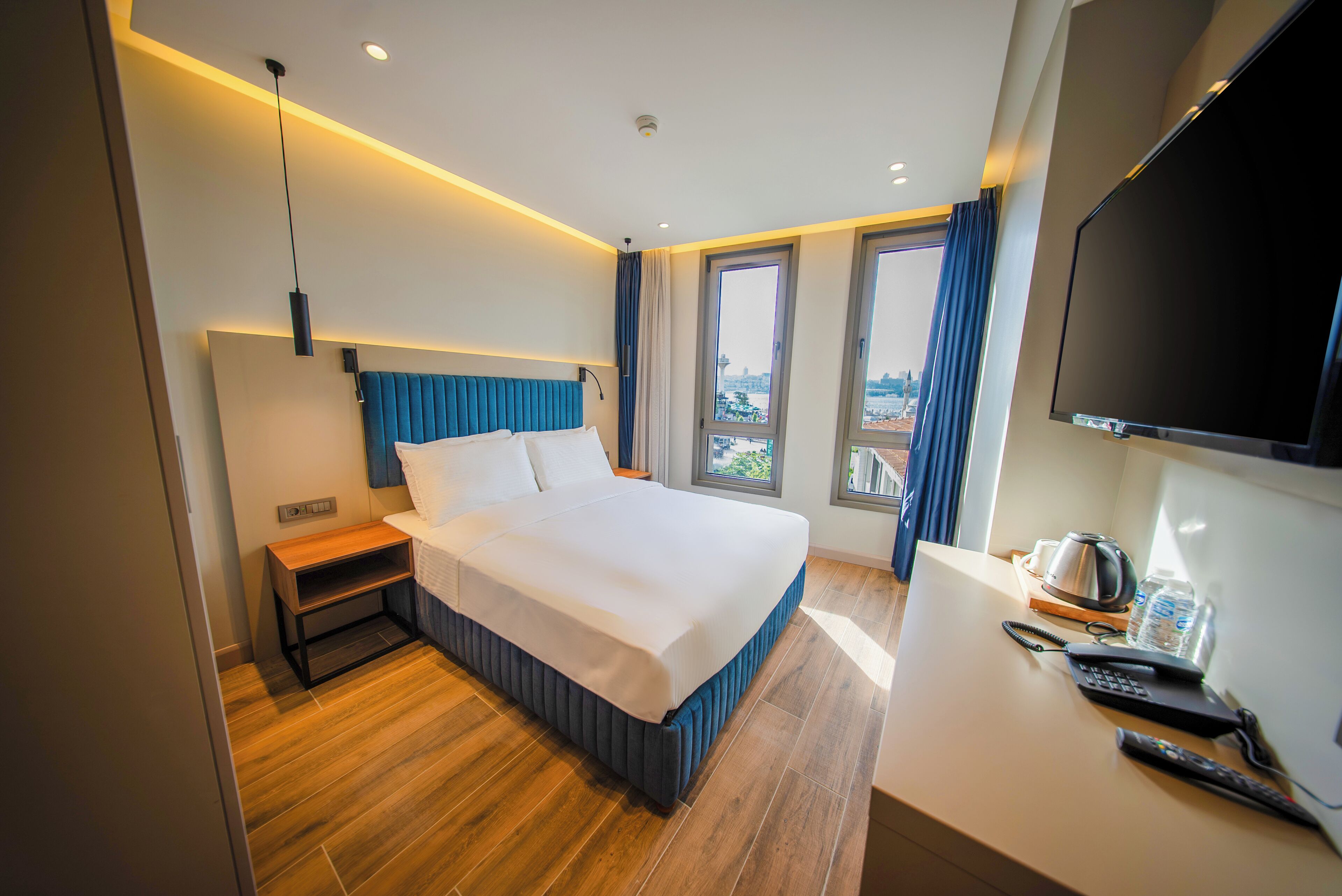 A modern hotel room with a large bed, a desk with a kettle, and a TV.