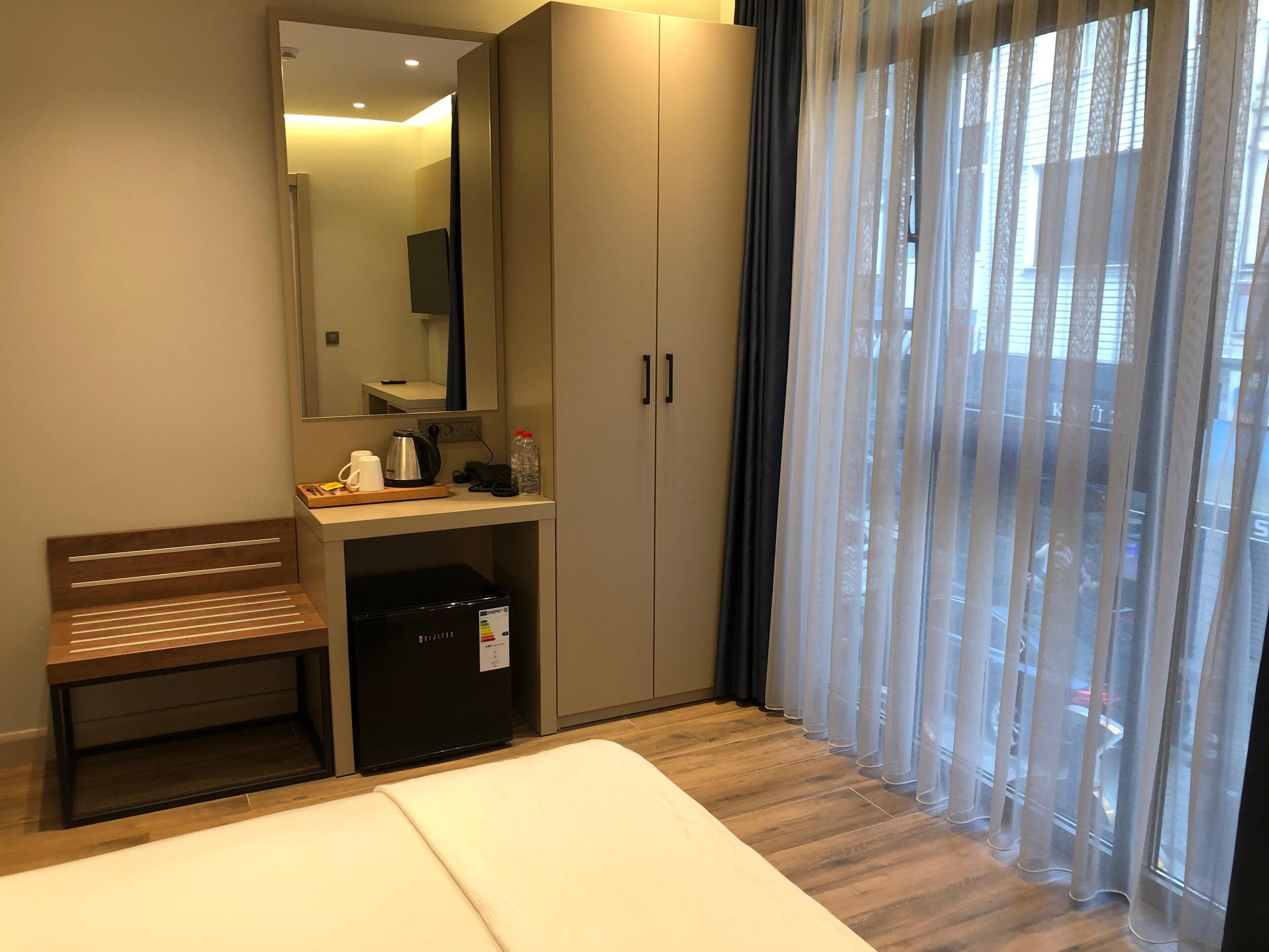 A hotel room with a bed, a desk with a kettle, a refrigerator, a mirror, and a window with sheer curtains.