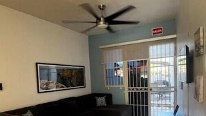 Un soggiorno con un divano ad angolo nero, una ventola a soffitto, una TV a muro e un pavimento piastrellato.