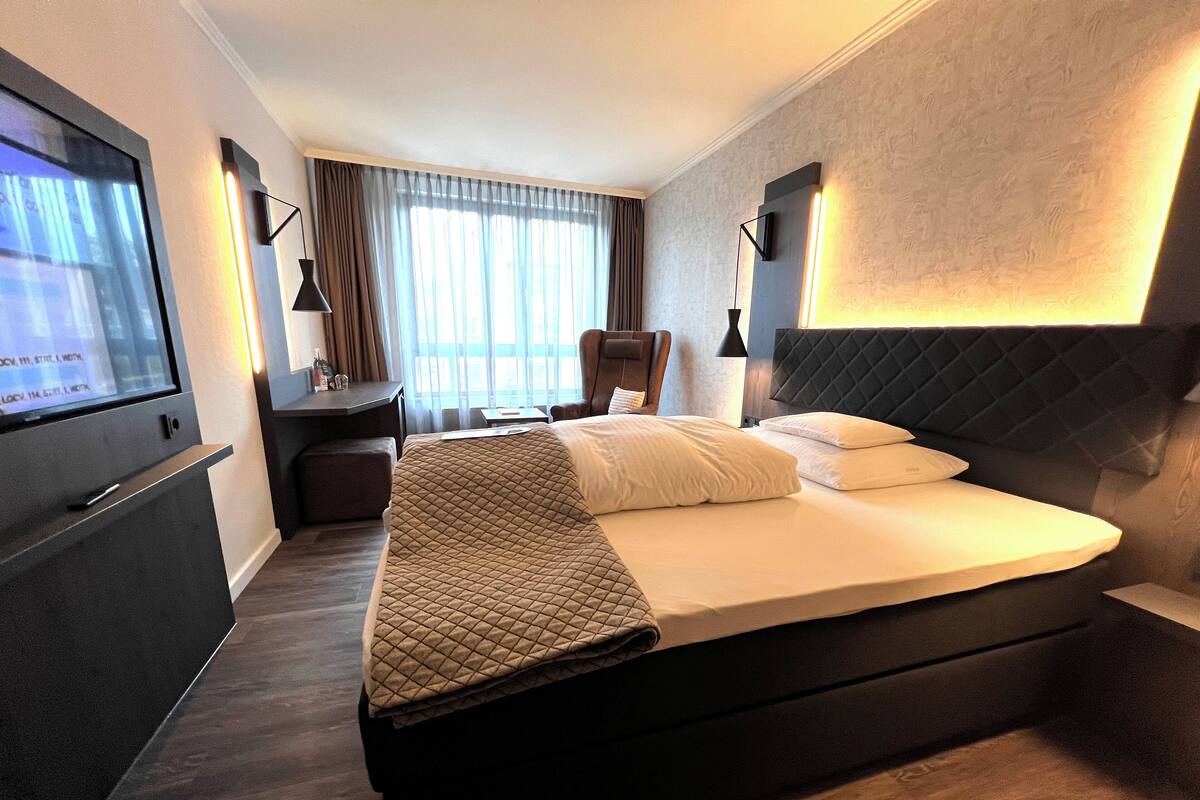 A modern hotel room with a large bed, a desk, a chair, and a TV on the wall.