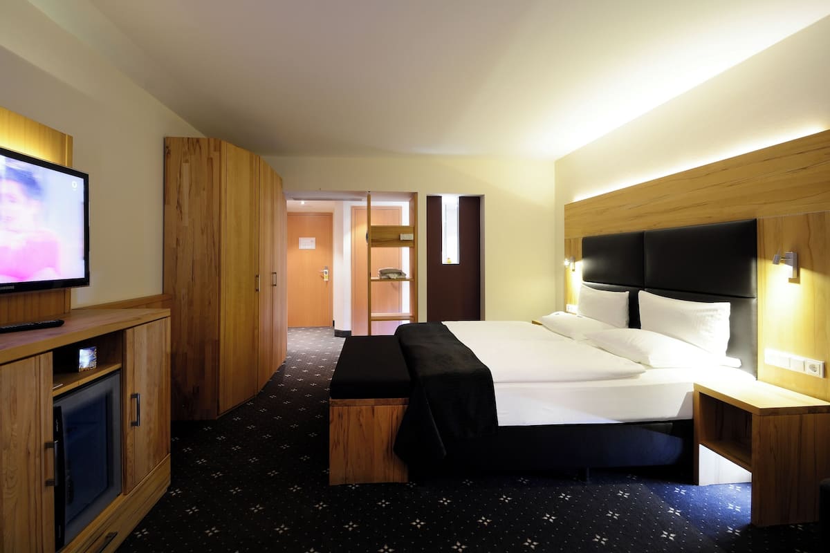 A hotel room with a bed, bedside table, TV, wooden wardrobe, and a bench.