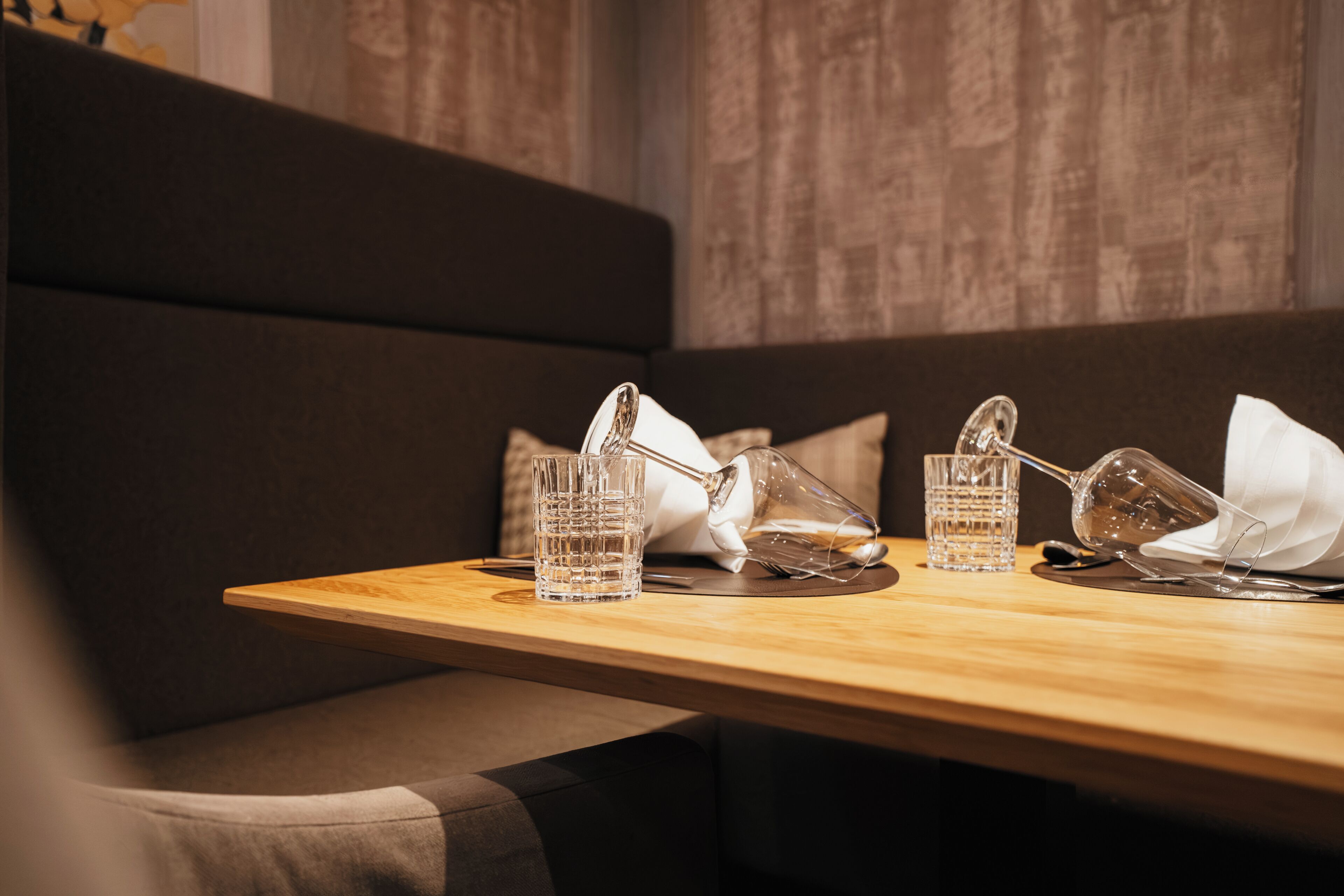 Een eettafel met helder glaswerk en servetten, voor een donkere gestoffeerde bank en een textuurrijke muur.