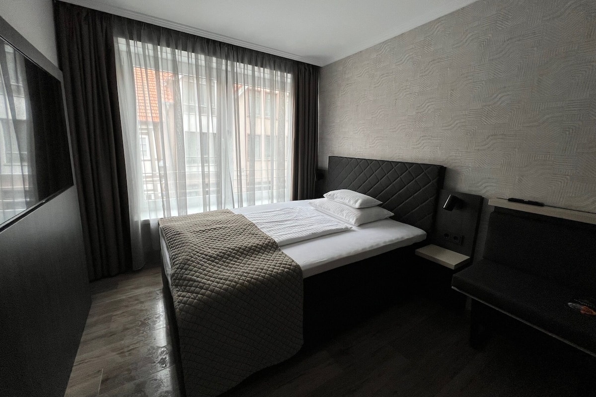 A modern hotel room with a bed, a bedside table, a TV, and a window with curtains.
