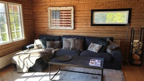 A cozy cabin interior with a wooden interior, a sofa with multiple cushions, a glass coffee table, a framed American flag, and a window with a view of trees.