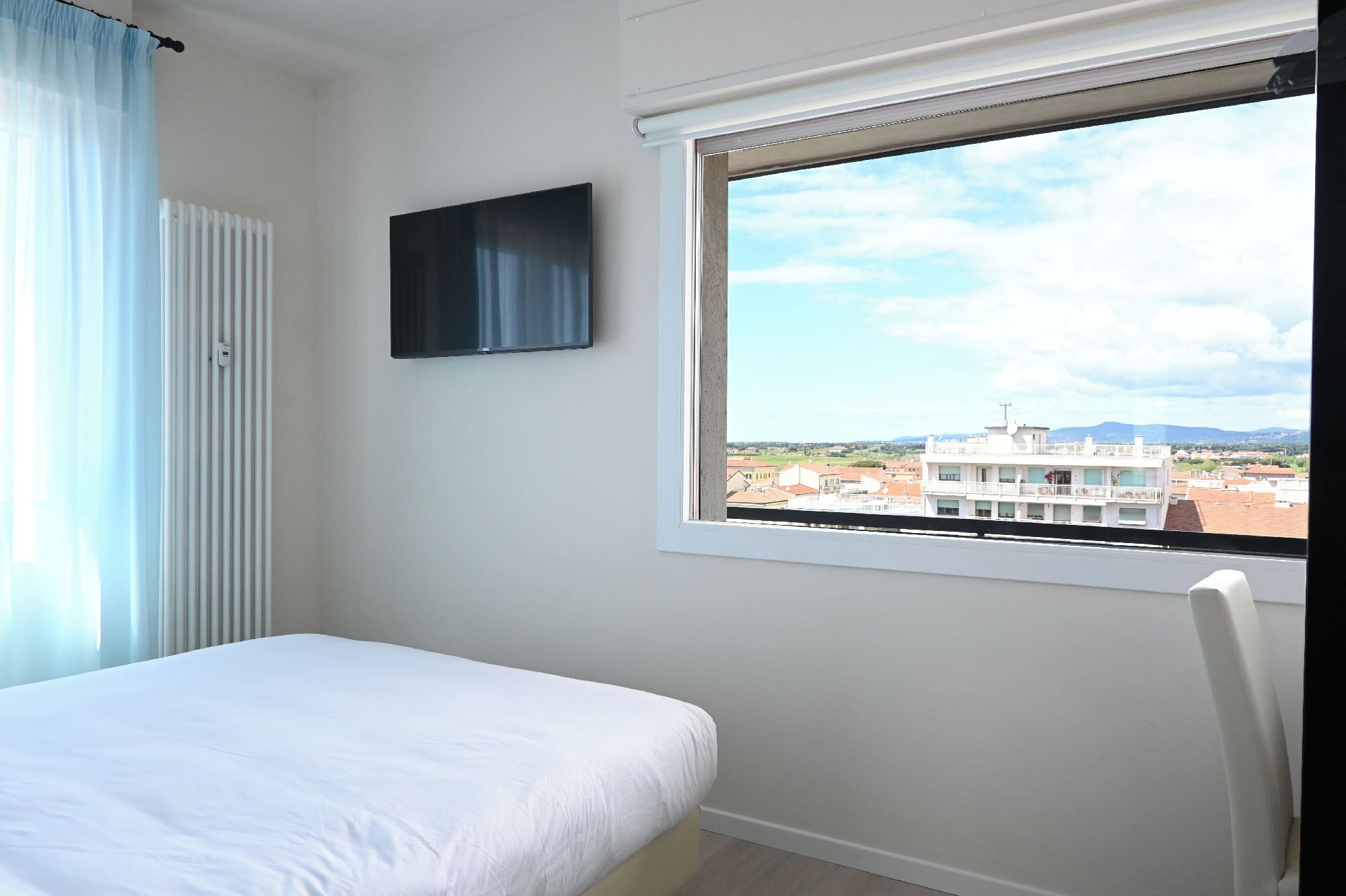 A bedroom with a bed, a window with a view of buildings, a radiator, and a TV on the wall.