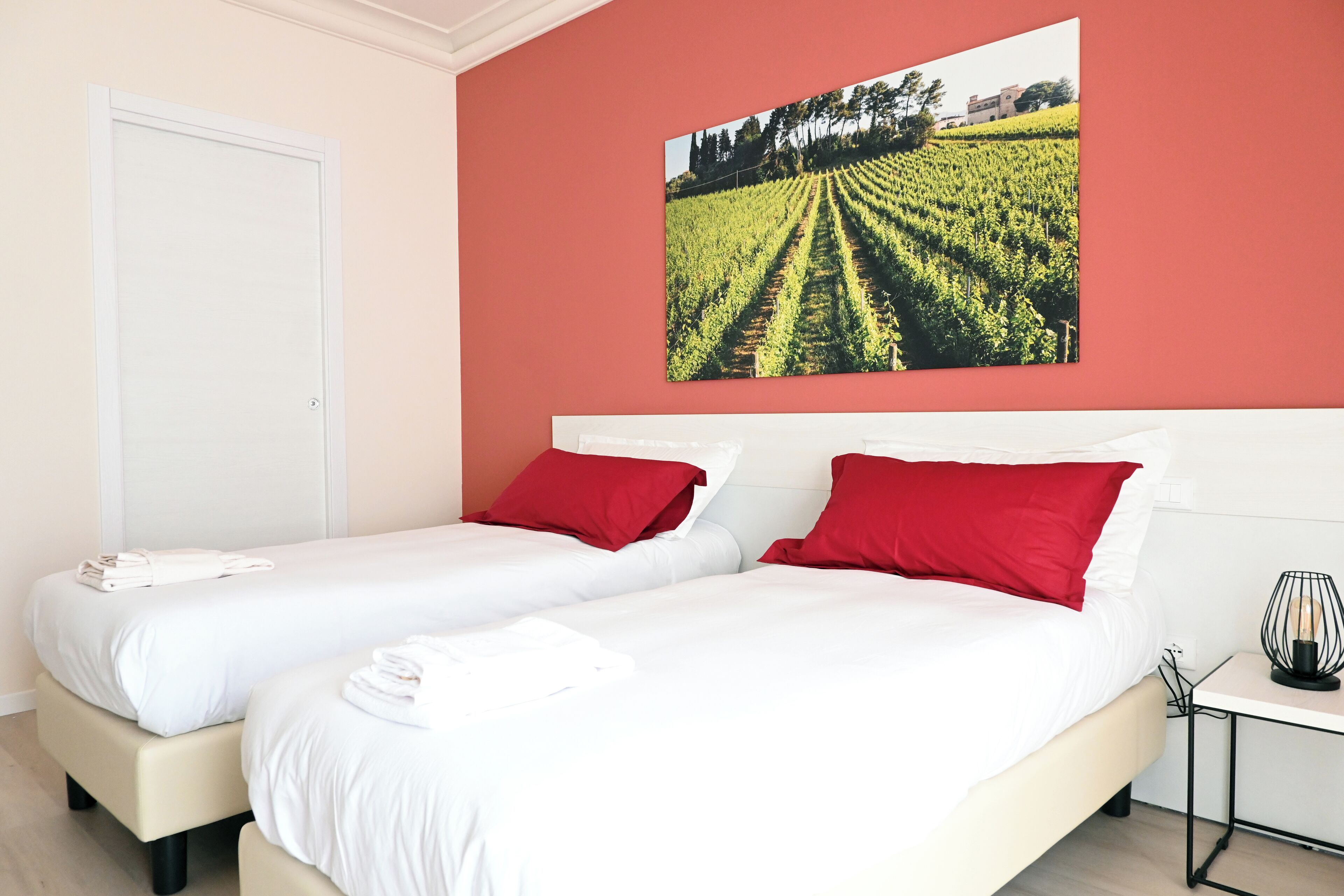 A hotel room with two beds, red pillows, a white headboard, a small table with a lamp, and a large vineyard painting on the wall.