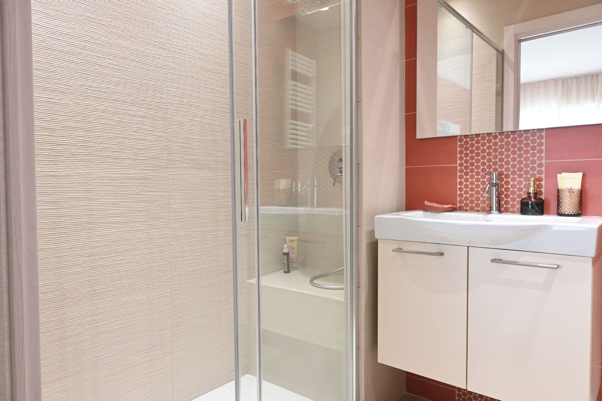 Un baño moderno con cabina de ducha de vidrio, un lavamanos blanco con un espejo encima y una pared de azulejos con estampado de lunares.