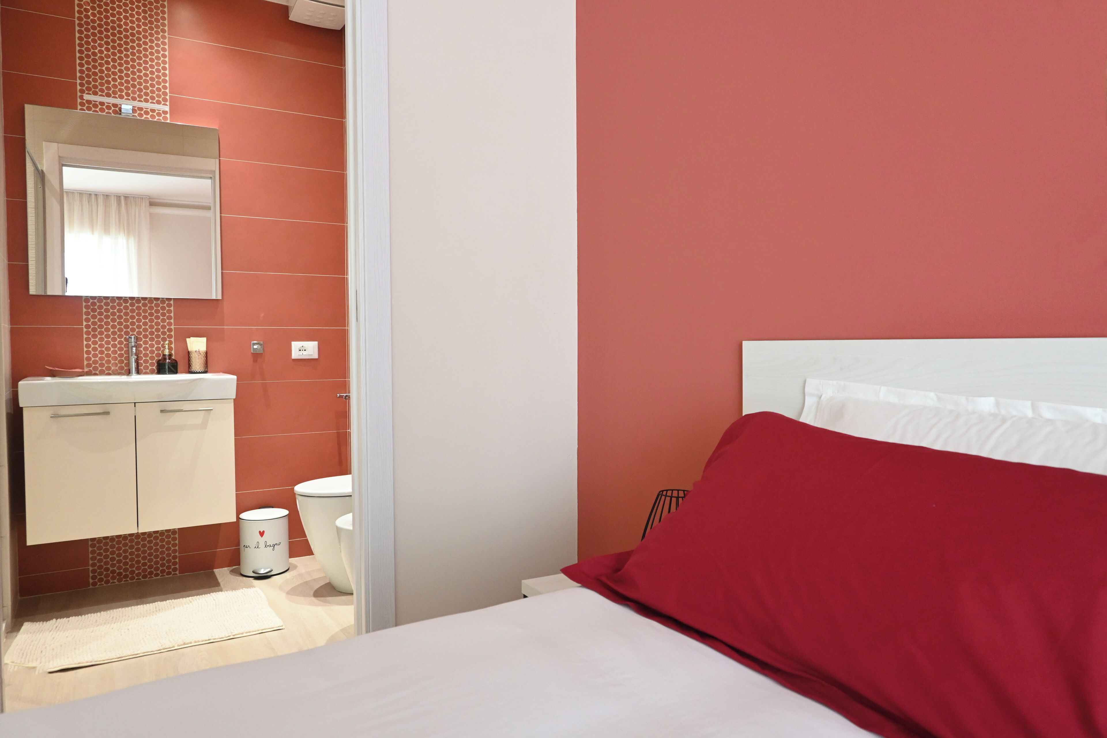 A hotel room with a bed, a white headboard, a red pillow, a white nightstand, a white bedside table, a white wall, a white door, a white bathroom with a white sink, a white toilet, a white mirror, a white towel, and a white rug.