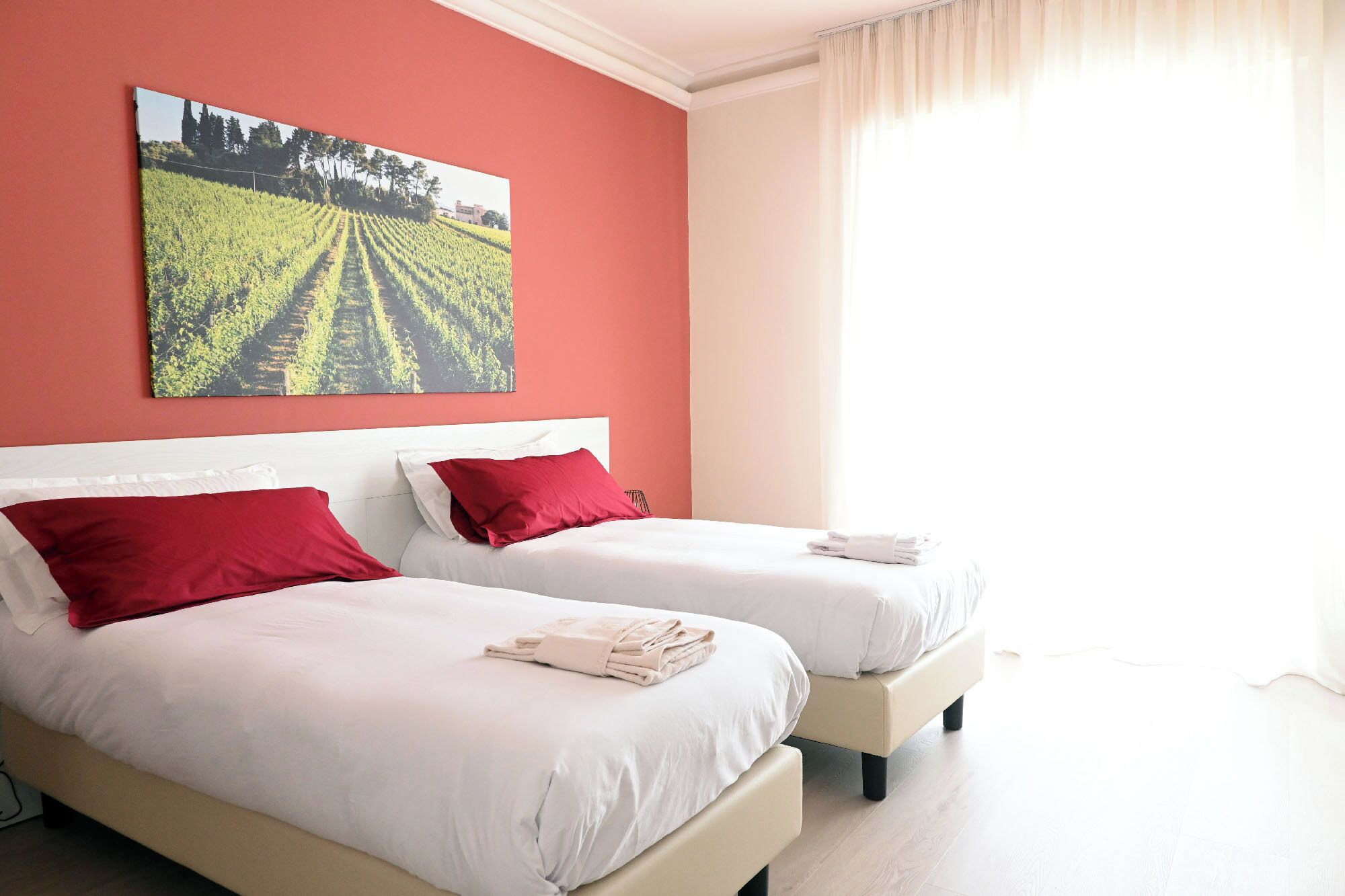 Quarto de hotel com duas camas, almofadas vermelhas, um quadro de paisagem grande na parede e uma janela com cortinas.