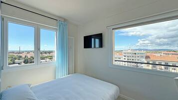 Ein Schlafzimmer mit Bett, zwei Fenstern, einem Fernseher und Blick auf eine Stadtlandschaft.