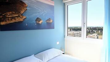 A bedroom with a large window, a bed with white bedding, and a painting of a coastal scene.