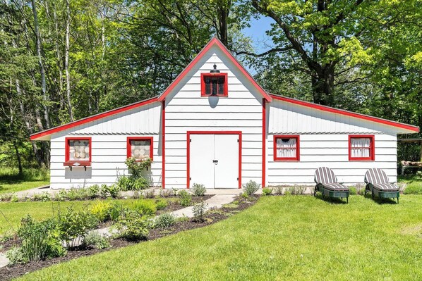 A white barn with red trim and a triangular roof, surrounded by greenery.