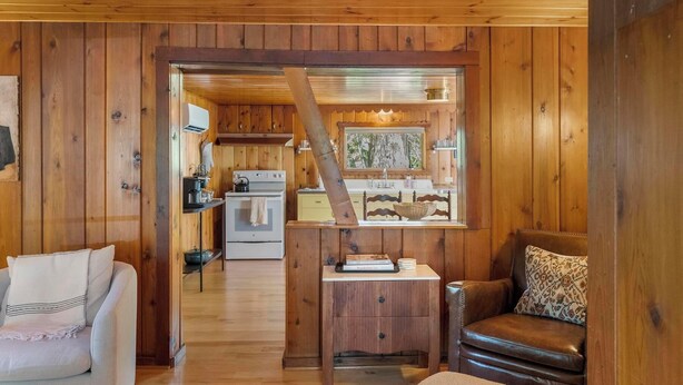 Interior acolhedor de uma cabana com paredes de madeira, área de cozinha, mesa de jantar com livros e uma poltrona de couro.