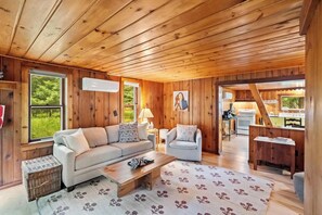 A cozy cabin interior with a wooden ceiling, a sofa, a coffee table, and a view of the outdoors through large windows.