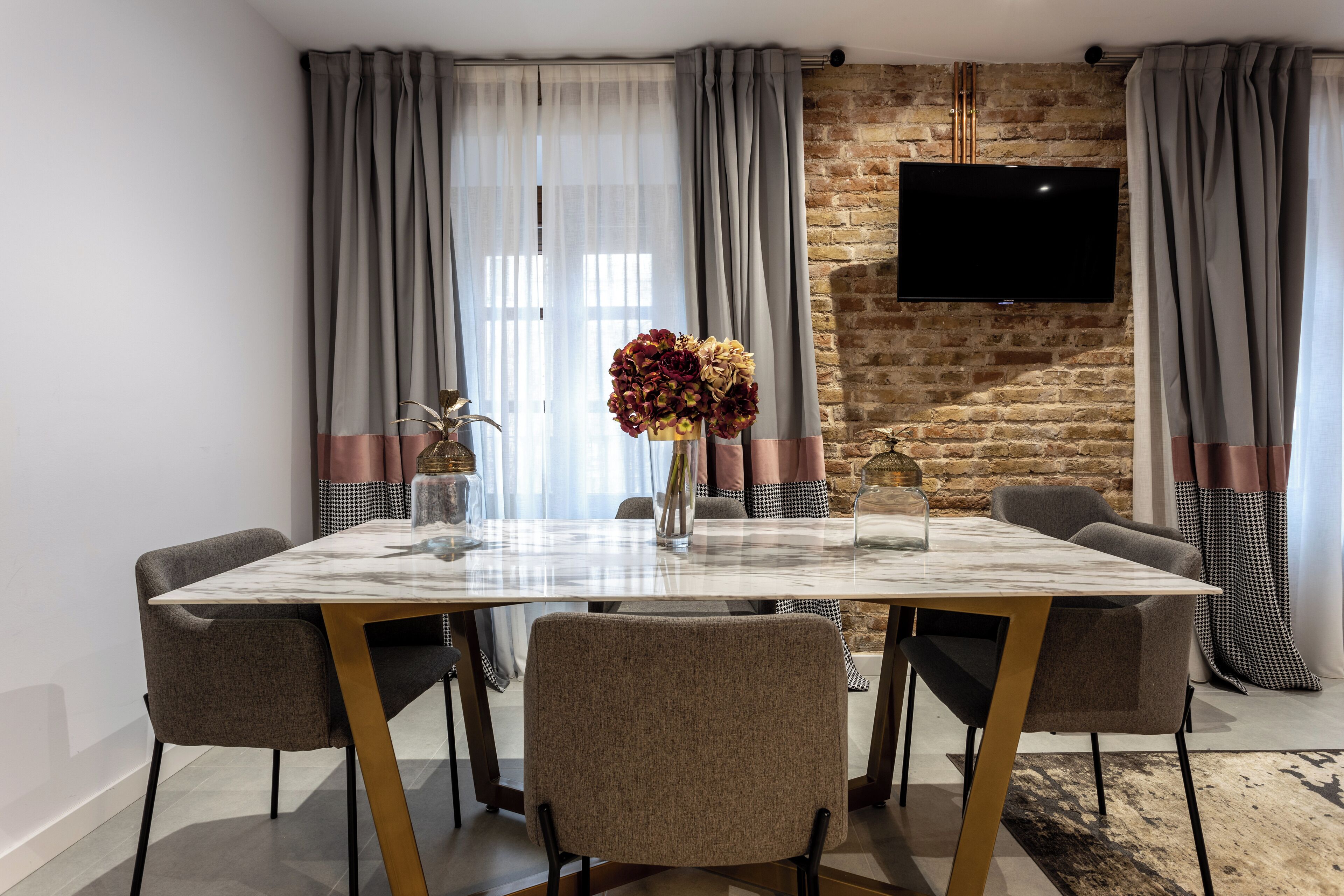 A dining area with a marble table, six chairs, a vase of flowers, and a flat-screen TV mounted on a brick wall.