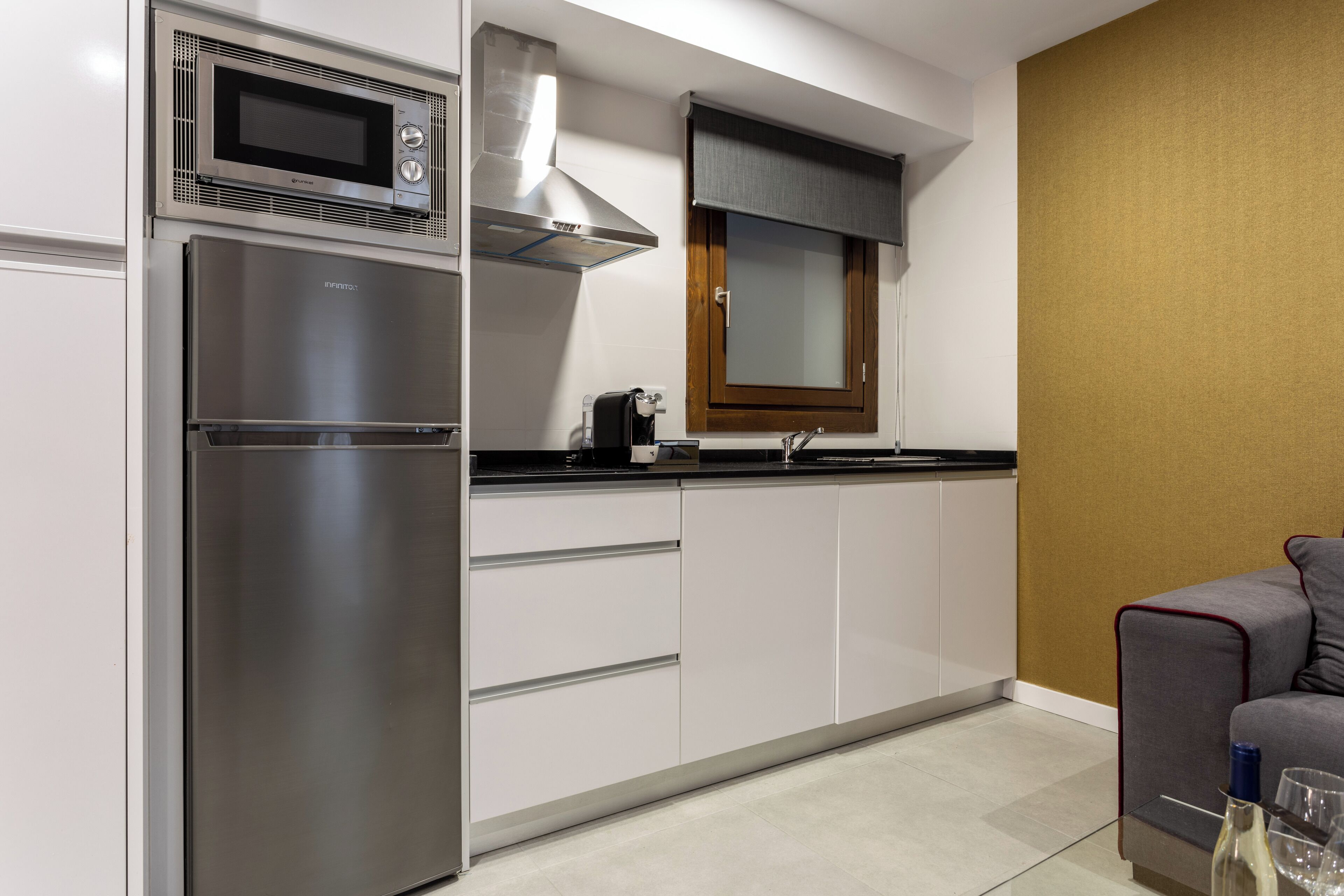 Cocina compacta con microondas integrado, horno y cocina a gas. Hay una ventana con persiana, un lavamanos y una pequeña encimera.