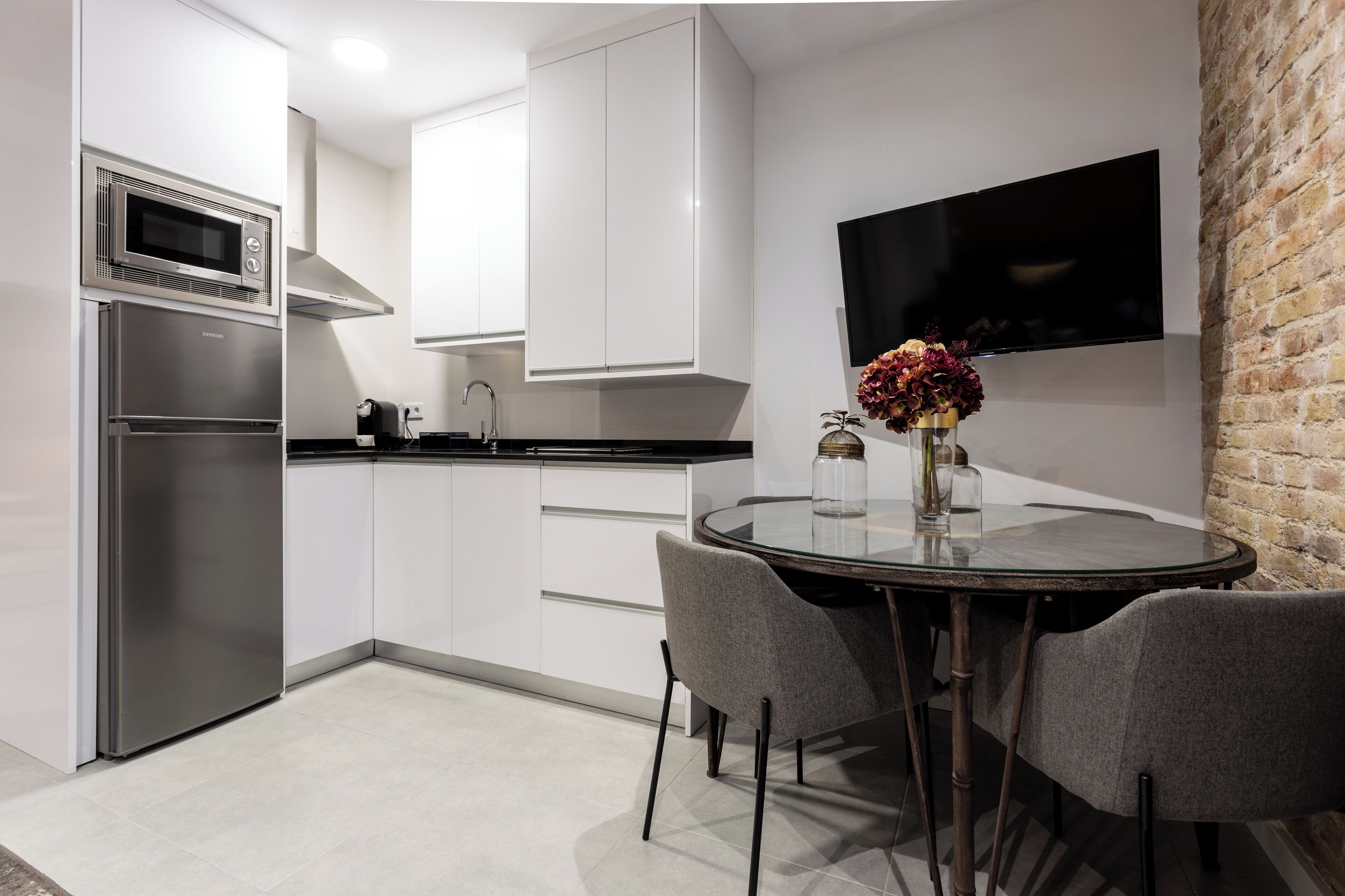 A modern kitchen with white cabinets, a stainless steel refrigerator, a microwave, and a flat-screen TV mounted on a brick wall.