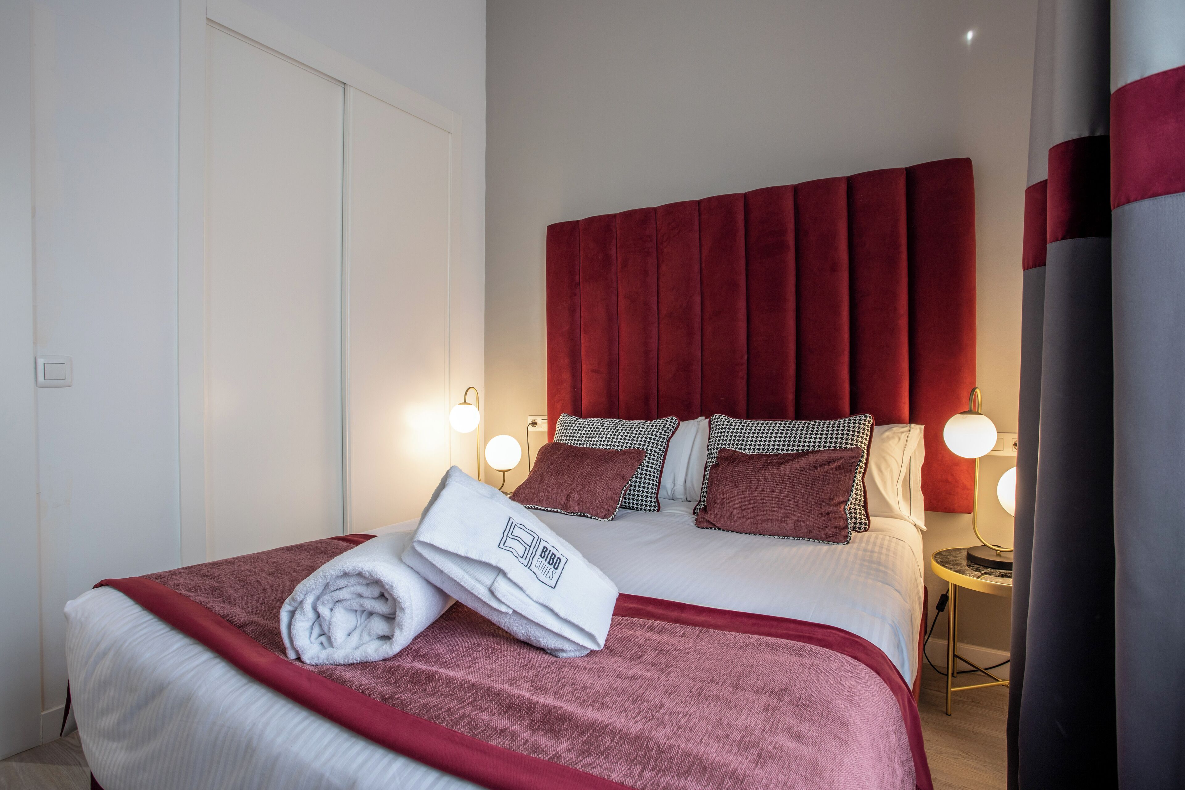 A neatly made bed with a red headboard, white bedding, and a folded towel on the side. There are two bedside lamps and a small table with a lamp.