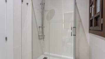 Une douche fermée par des parois en verre, dotée d’un pommeau de douche à main, d’un robinet mural et d’une petite étagère.