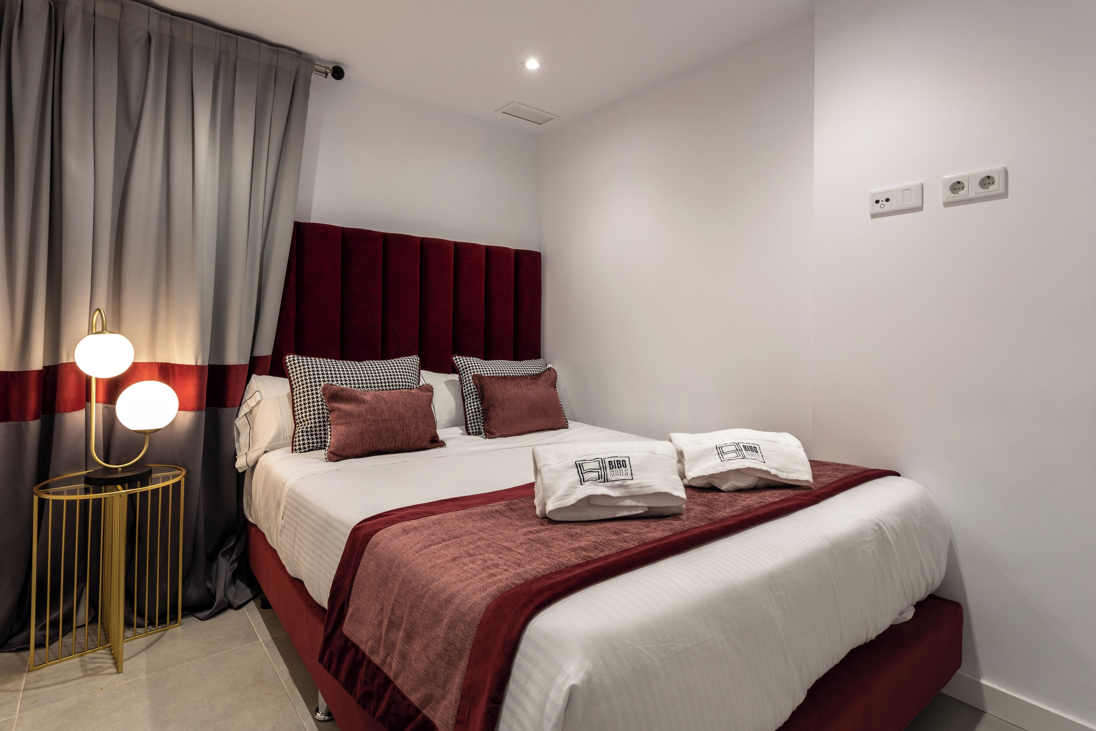 Un lit bien fait, avec une tête de lit rouge, une lampe de chevet et un oreiller à carreaux.
