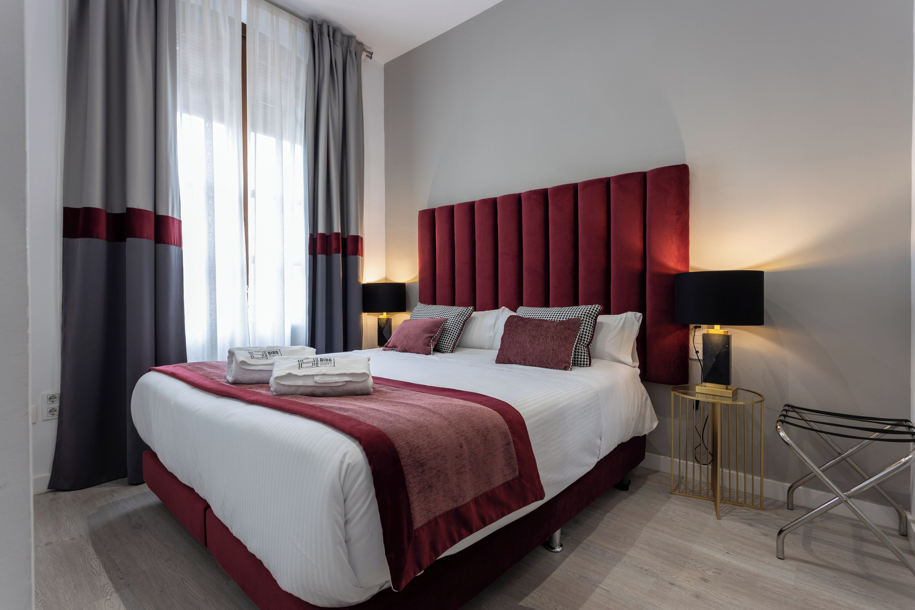Une chambre d’hôtel moderne avec un grand lit, une literie rouge et blanche, une tête de lit rouge, deux lampes de chevet, une petite table d’appoint et un porte-serviettes fixé au mur.