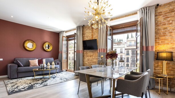 A modern living room with a sofa, a dining table, and a TV mounted on a brick wall. - BIBO SUITES PLAZA NUEVA (Granada)
