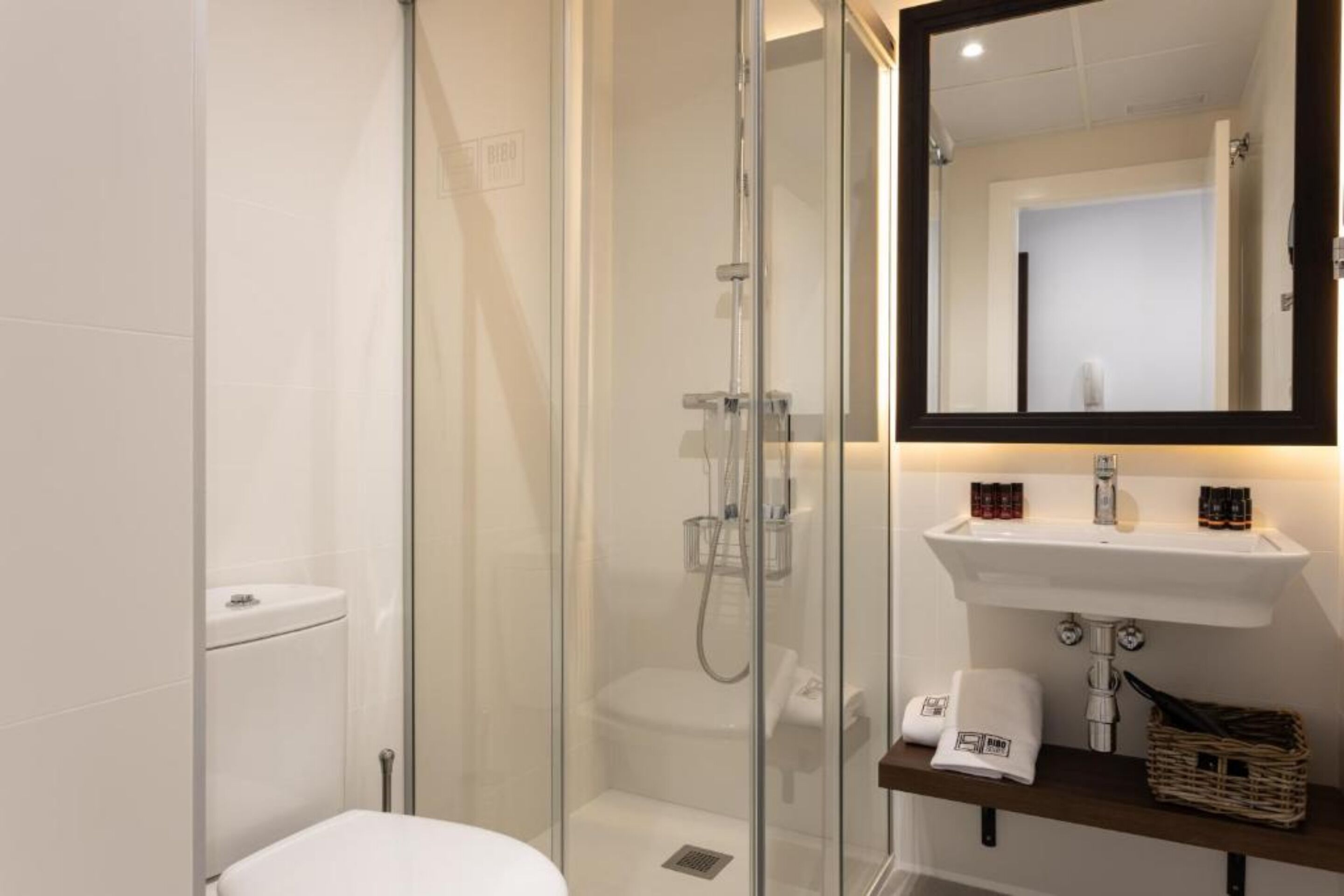 A modern bathroom with a glass-enclosed shower, a white toilet, a sink with a mirror above, and toiletries on the sink.