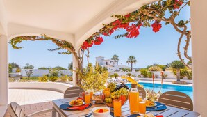 Mesa de comedor dispuesta con alimentos para el desayuno y vista a una piscina y edificios.