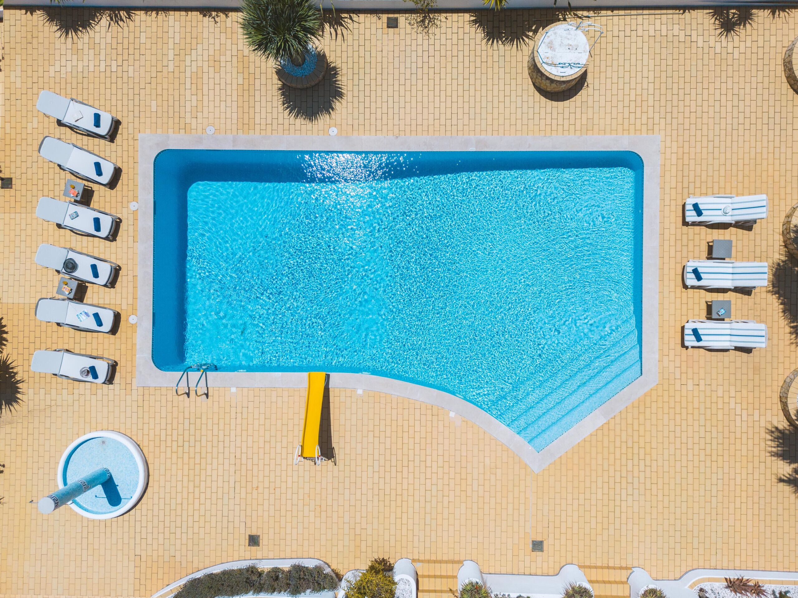 An aerial view of a rectangular swimming pool surrounded by lounge chairs and sunbeds, with a brick wall and a palm tree in the background.