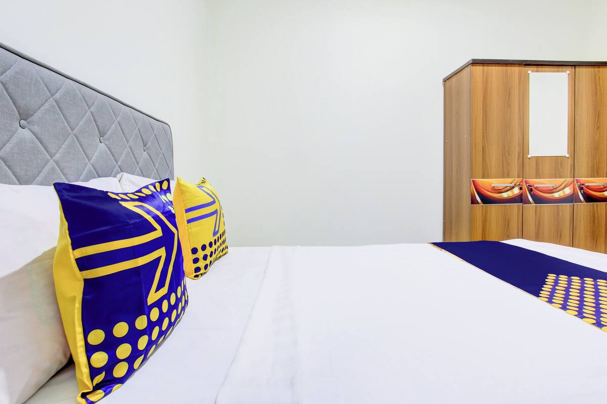 Una cama bien tendida con edredón blanco y almohadas azules y amarillas, un cabecero gris con capitoné y un armario de madera con espejo.