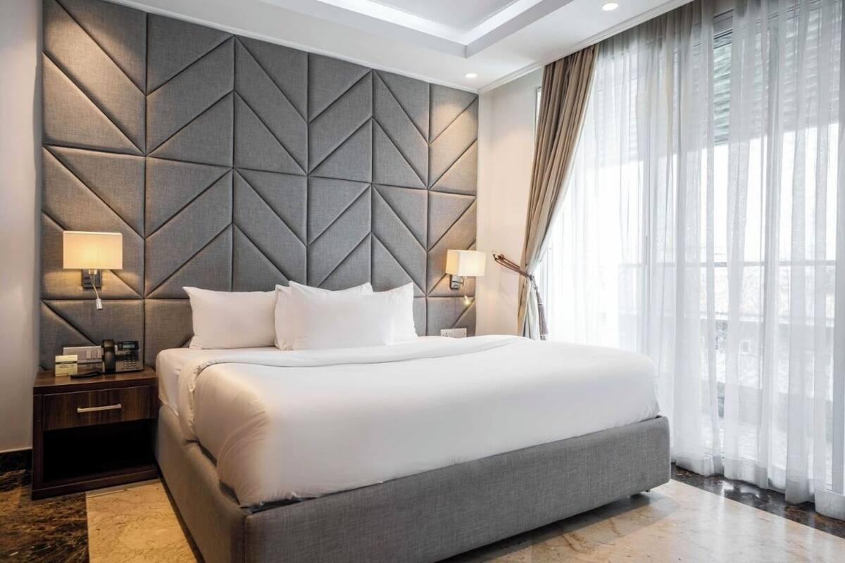A modern hotel room with a large bed, a geometric headboard, two bedside tables with lamps, and sheer curtains.