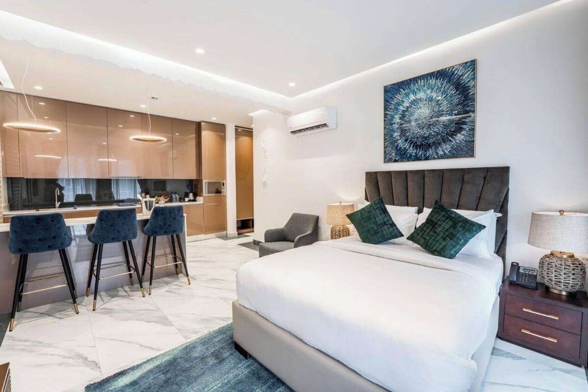 A modern hotel room with a large bed, a dining area with bar stools, and a kitchen in the background.