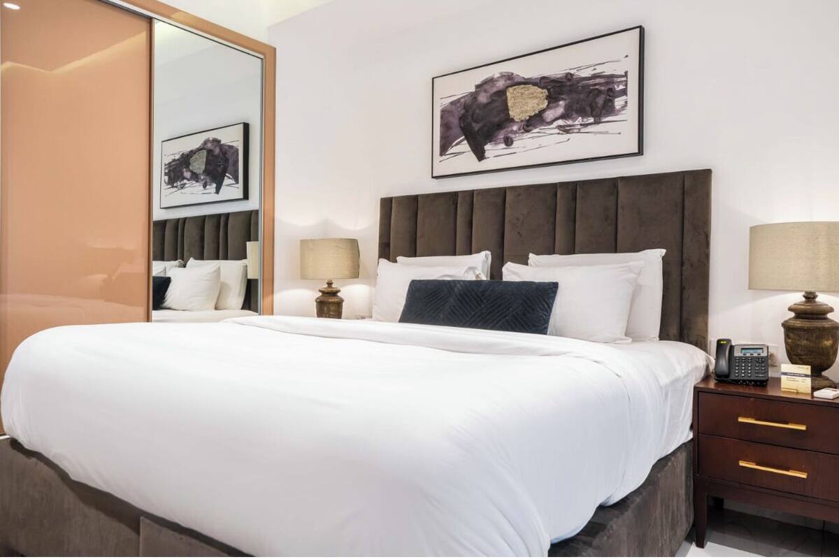 A hotel room with a large bed, a wooden headboard, two bedside tables with lamps, a mirror, and two framed artworks on the wall.