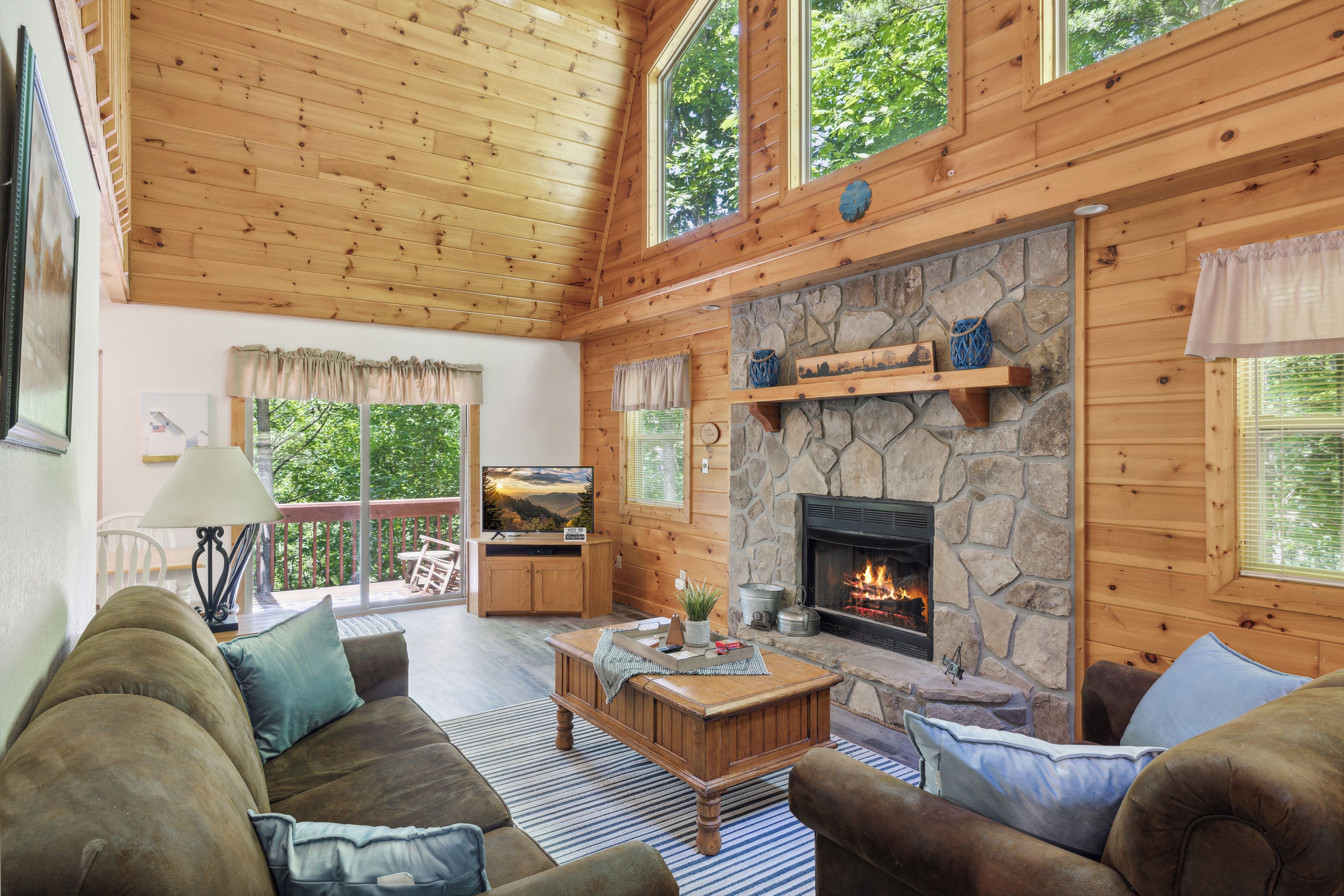 A cozy living room with a stone fireplace, wooden ceiling, and large windows.