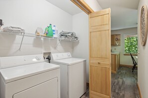 A laundry room with two white washing machines, shelves with folded towels and cleaning supplies, and a wooden door leading to another room. - Cosy Cabin|King Suite main floor|Game Room|Hot Tub (Gatlinburg)