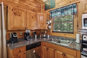 Une cuisine avec des meubles en bois, un plan de travail en granit, une machine à café, un lave-vaisselle et une fenêtre donnant sur un paysage verdoyant.