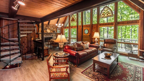A spacious living area with a wooden staircase, leather sofa, and large windows offering a view of the forest. - Woodland Lodge - Perfect Family Ski Chalet! Close to Nubs Nob & Boyne! (Harbor Springs)