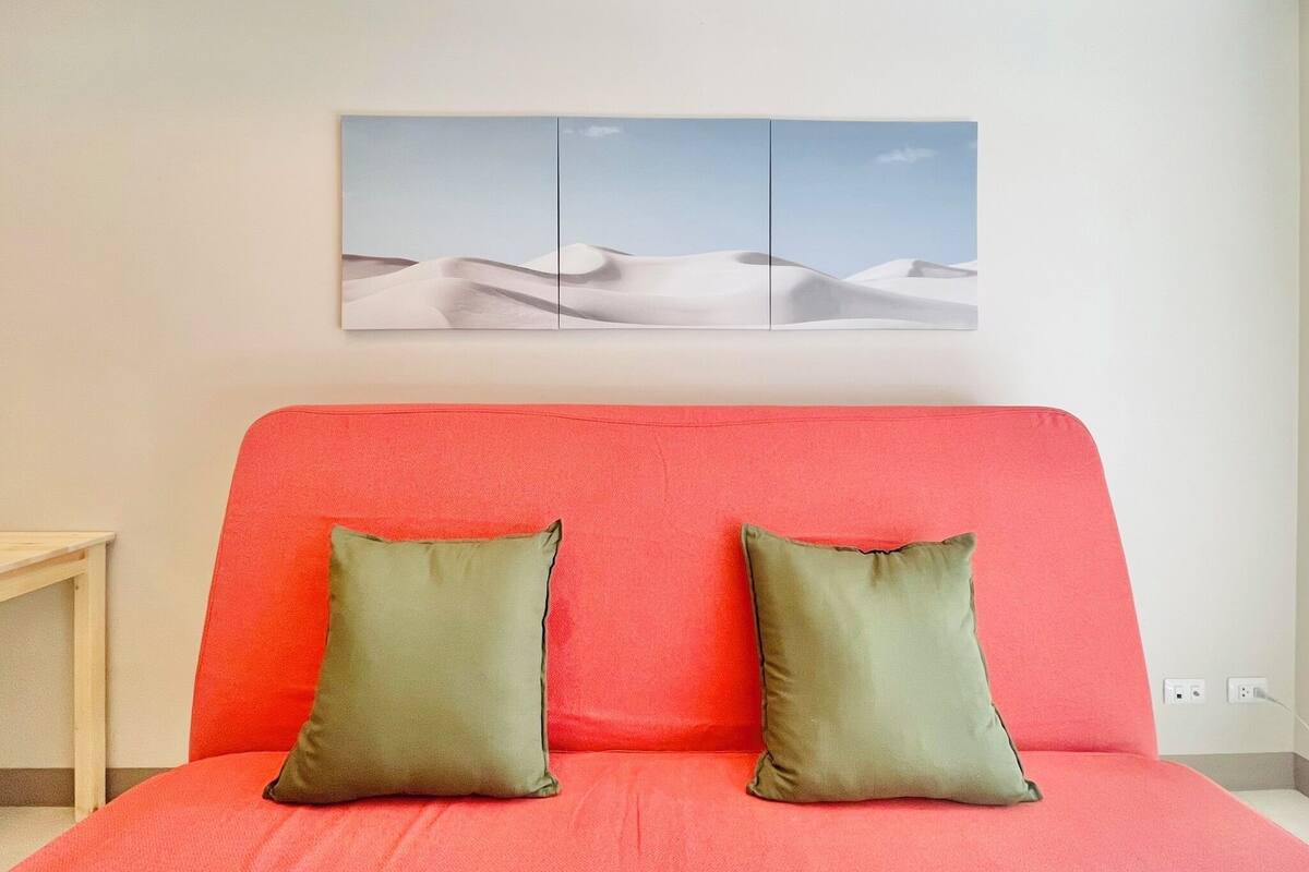 A room with a red bed, two green pillows, and a wall-mounted picture of sand dunes.