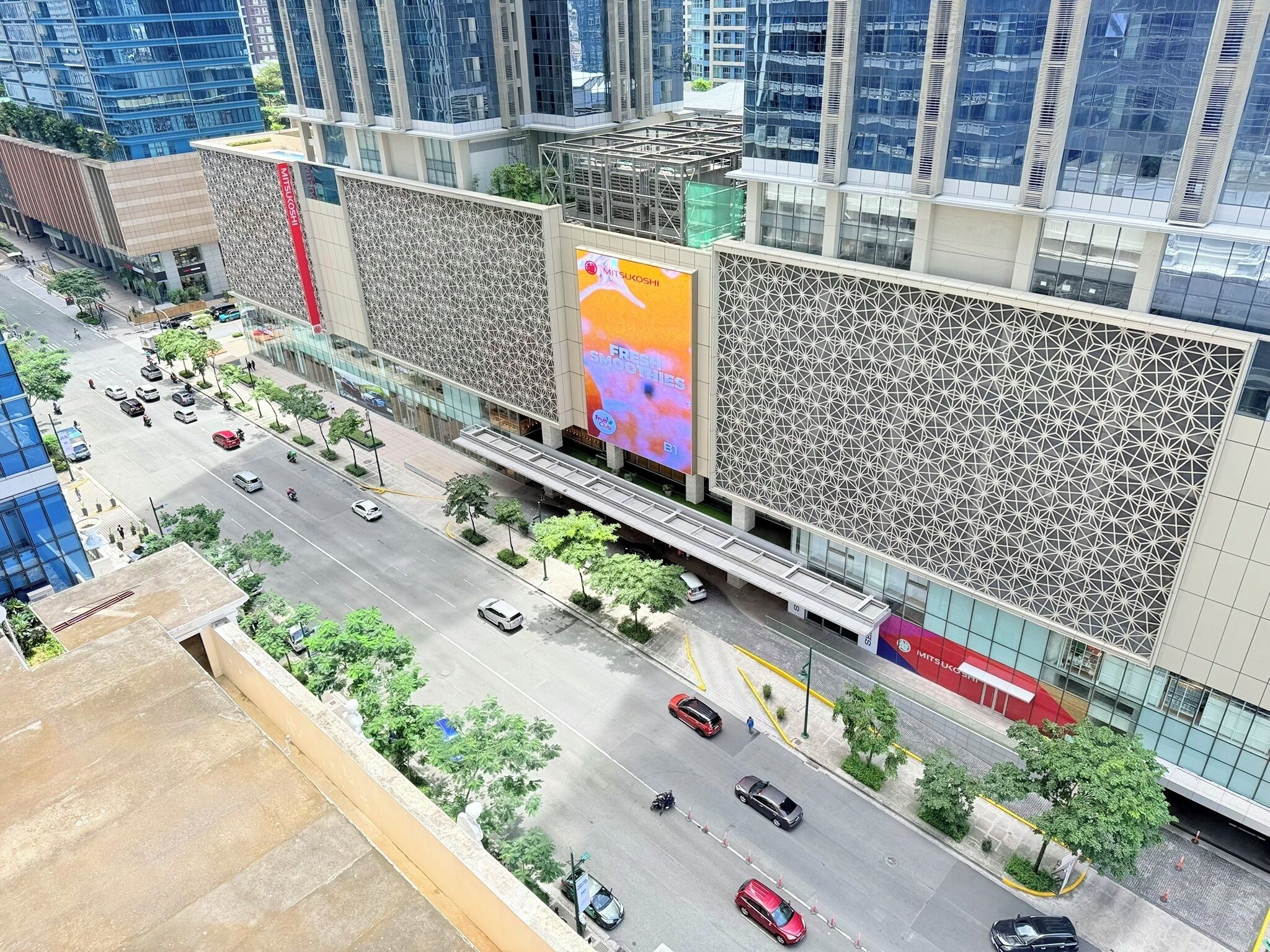 Un bâtiment moderne, doté d’un grand panneau publicitaire, d’un passage piétonnier et situé dans une rue animée, bordée d’arbres et fréquentée par de nombreuses voitures.