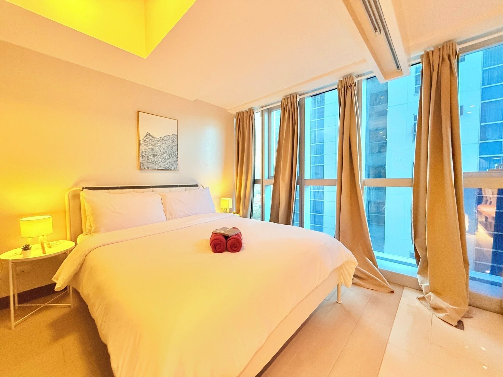 A hotel room with a large bed, a nightstand with a lamp, a painting on the wall, and a view of the city through large windows.