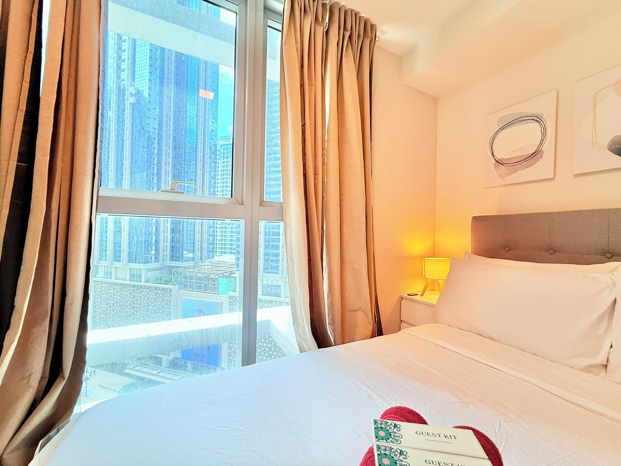 A hotel room with a large window, a bed, a nightstand with a lamp, and a wall with a framed picture.