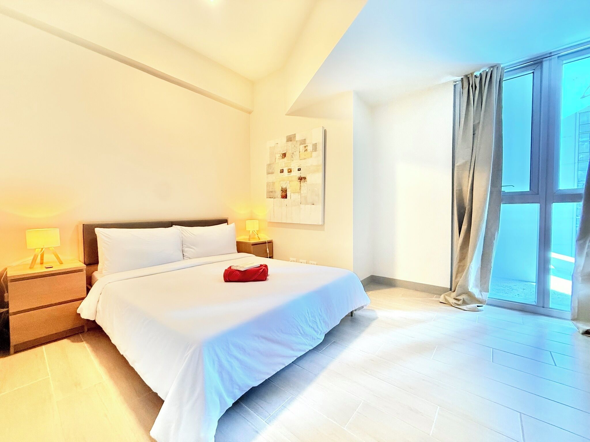 A hotel room with a large bed, two bedside tables with lamps, a window with curtains, and a wall-mounted artwork.