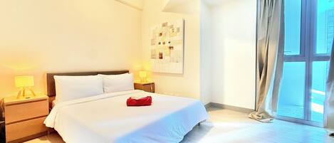 A hotel room with a large bed, two bedside tables with lamps, a window with curtains, and a wall-mounted artwork.
